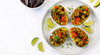 Overhead view of four avocado halves stuffed with a colorful vegan chorizo grain salad, garnished with black beans, corn, red peppers, cilantro, and lime wedges on a white plate with blue tortilla chips on the side.
