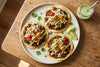 Vegan chorizo tostadas on a ceramic plate, layered with refried beans, crumbled plant-based chorizo, shredded cabbage, cherry tomatoes and red onion, drizzled with cilantro lime crema, with a jar of crema and lime wedges alongside